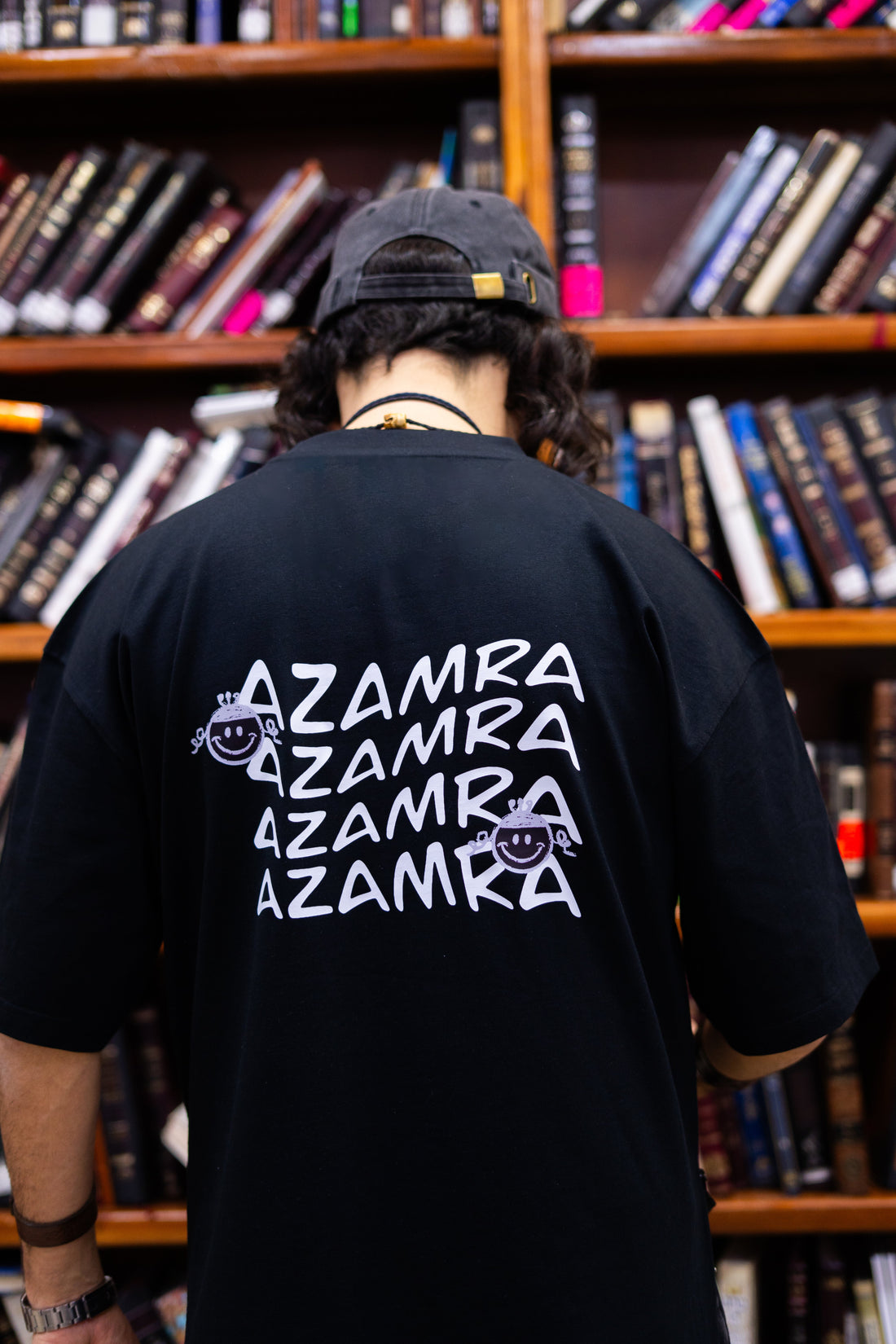 Un homme portant un T-shirt tsitsit AZAMRA devant une bibliothèque de livres religieux – inspiration Rabbi Nahman de Breslev